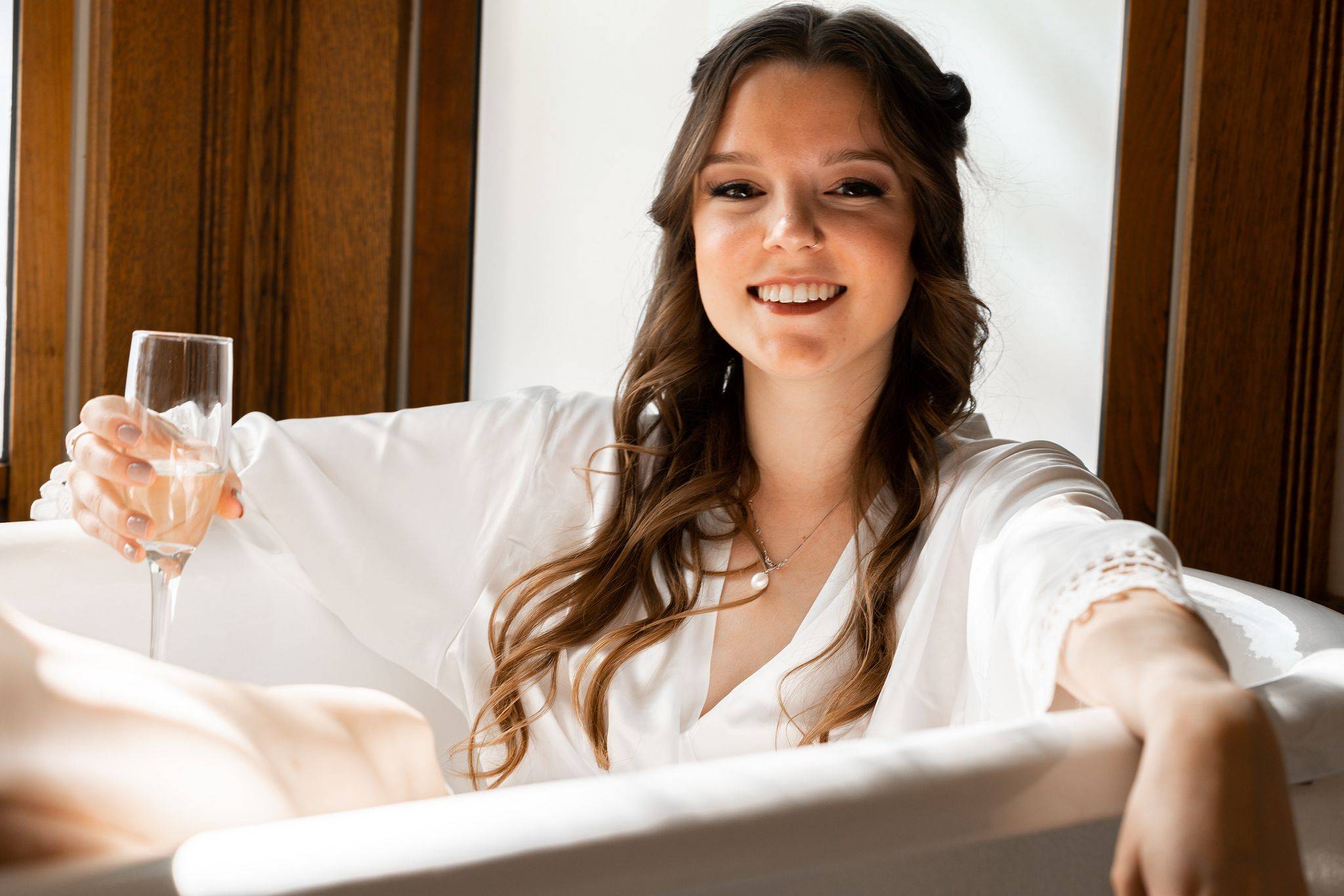 bride with champagne glass and bridesmaid robes sitting in a bath tub.