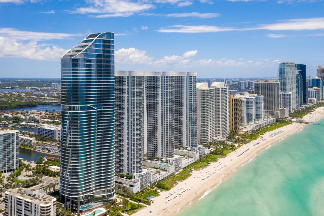 Aerial drone shot of Sunny Isles Beach city coastline with modern towers, beach, bay blue sky with clouds in the background, cityscape, swimming pools, turquoise sea