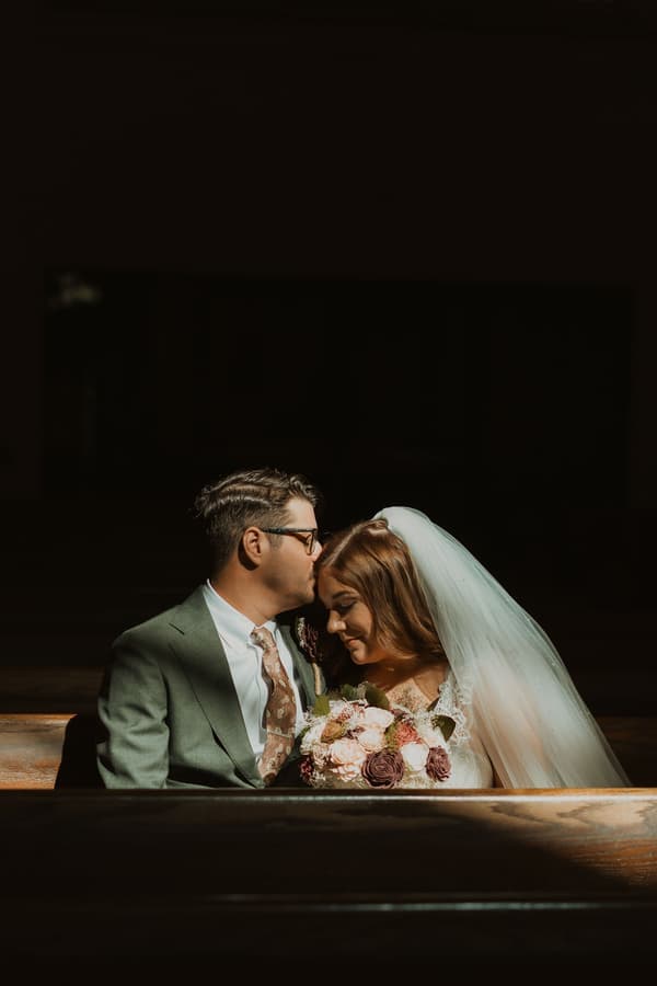 A tender moment between a bride and groom in a dimly lit church pew, bathed in soft golden light.