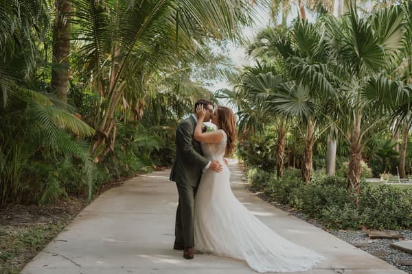 A bride and groom share a tender kiss under the lush canopy of palm trees in an outdoor garden setting.
