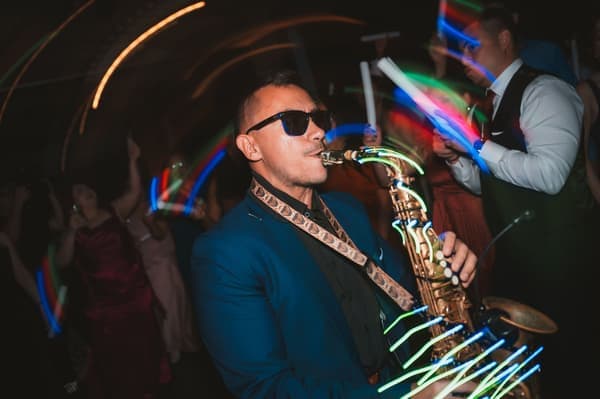 A saxophonist in a vibrant blue suit and sunglasses performs passionately at an evening wedding reception with neon lights swirling around him.