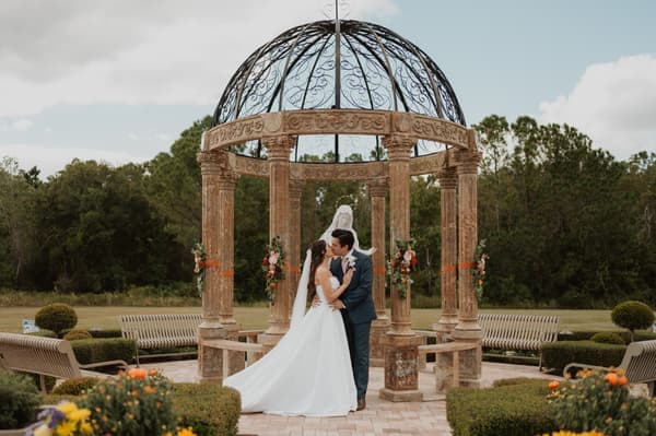 A man and a woman are standing under an archway with flowers on it. The man is wearing a blue suit and the woman is wearing a white dress. They are both looking at each other and smiling. The setting is outdoors in a garden with trees, bushes, and benches around them. The mood of the image is happy and romantic.