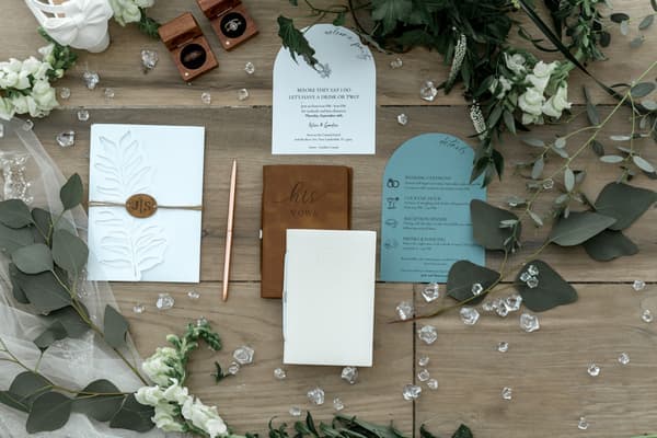 A wooden table with a bunch of wedding stationery on it including an invitation card, a guest list, place cards, a menu and a seating plan. There are also some white flowers scattered around the table. The setting is outdoors in springtime giving off a romantic mood.