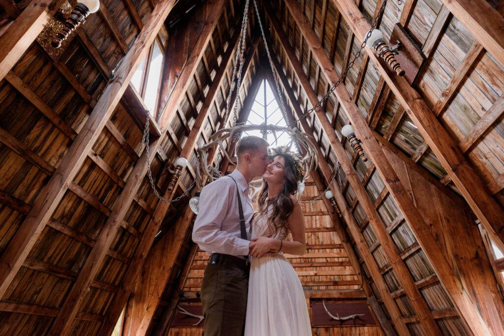 Boy is kissing a girl in cheek, dressed in wedding attire inside of a wooden building, couple in love